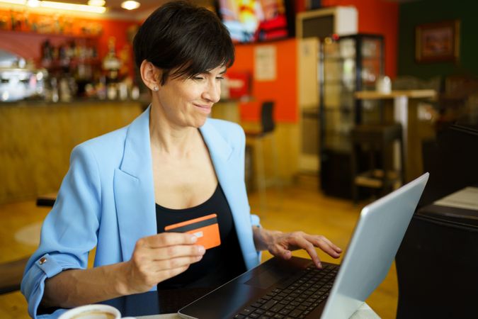 Smiling female in trendy blue jacket sitting in cafe with laptop and credit card