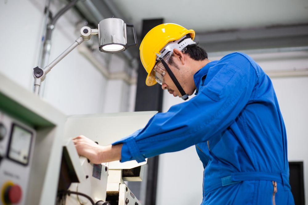 Side view of Asian man in blue jumpsuit and yellow hard hat working with machinery