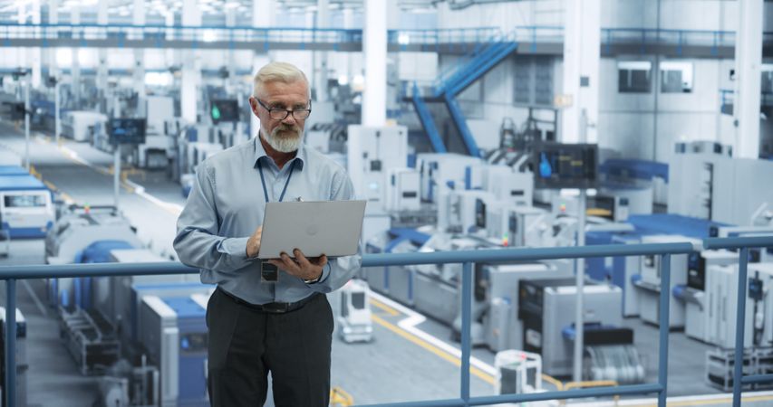Factory Manager Working on Laptop at a Futuristic Industrial Manufacture with Autonomous AI Robots. Specialist Monitoring All the Data on a Computer, Checking Reports and Planning Workload
