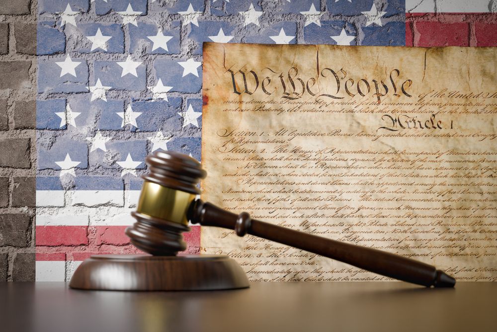 United States Constitution and American Flag On Wall with Gavel Resting on Table in Front.