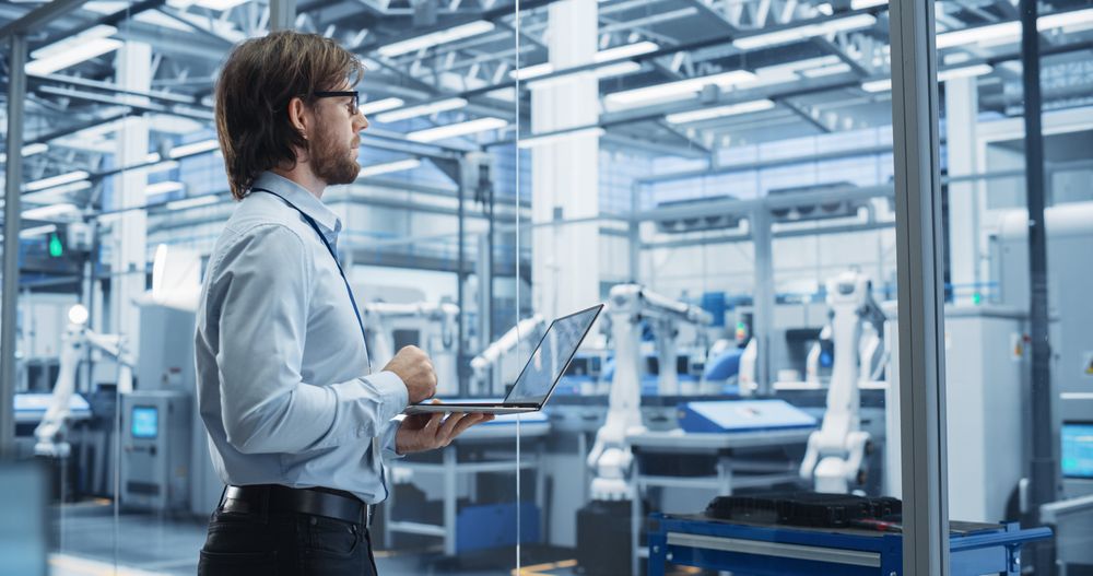 Factory Manager Working on Laptop at a Futuristic Industrial Manufacture with Autonomous Support Robots. Specialist Monitoring Technical Data on a Computer, Analyzing Reports and Planning Updates