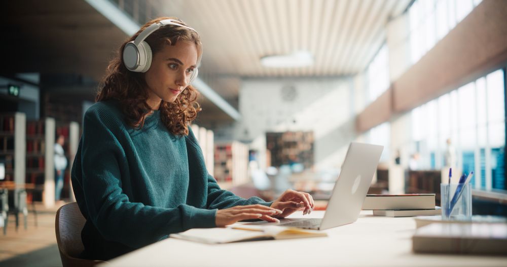 Concentrated Young Woman Studying in Modern Library, Wearing Headphones and Using Laptop