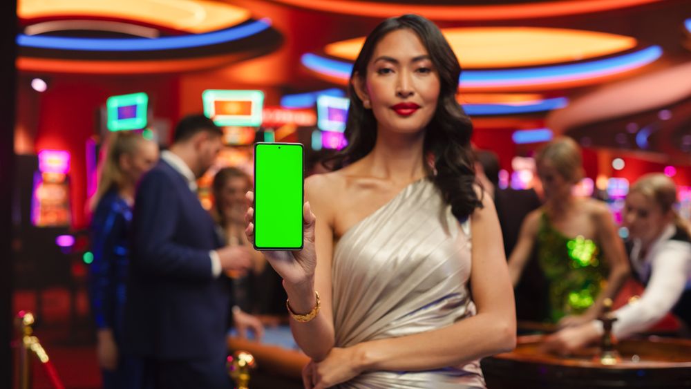 Modern Casino Setting: Gorgeous Asian Brunette Woman Posing with a Mobile Phone with a Green Screen Placeholder Space for Advertising