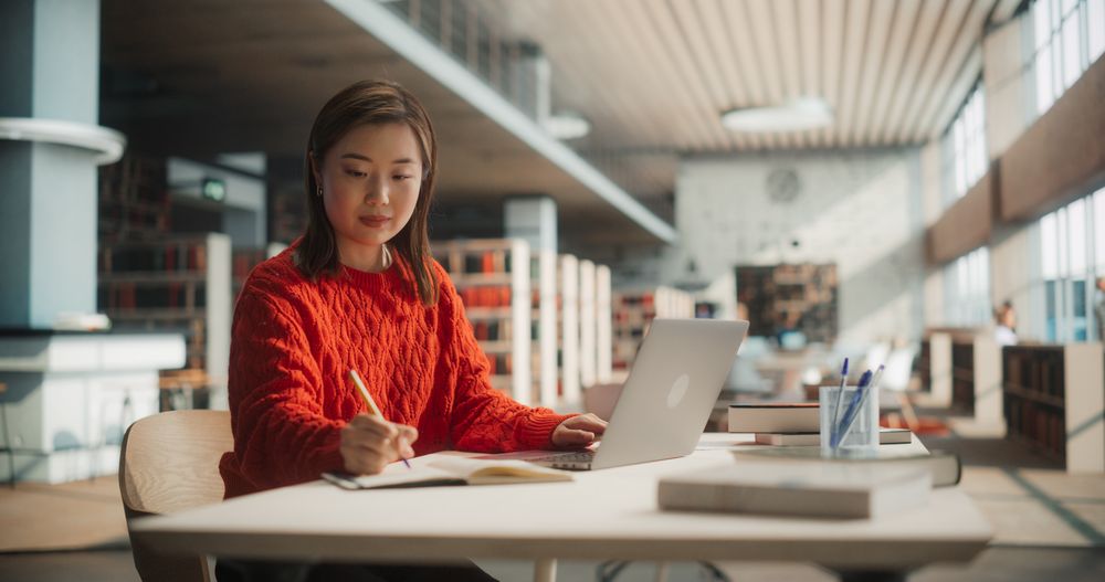 Diligent Asian Female Student Engaged in Academic Research With Laptop and Notebook in Modern Library.