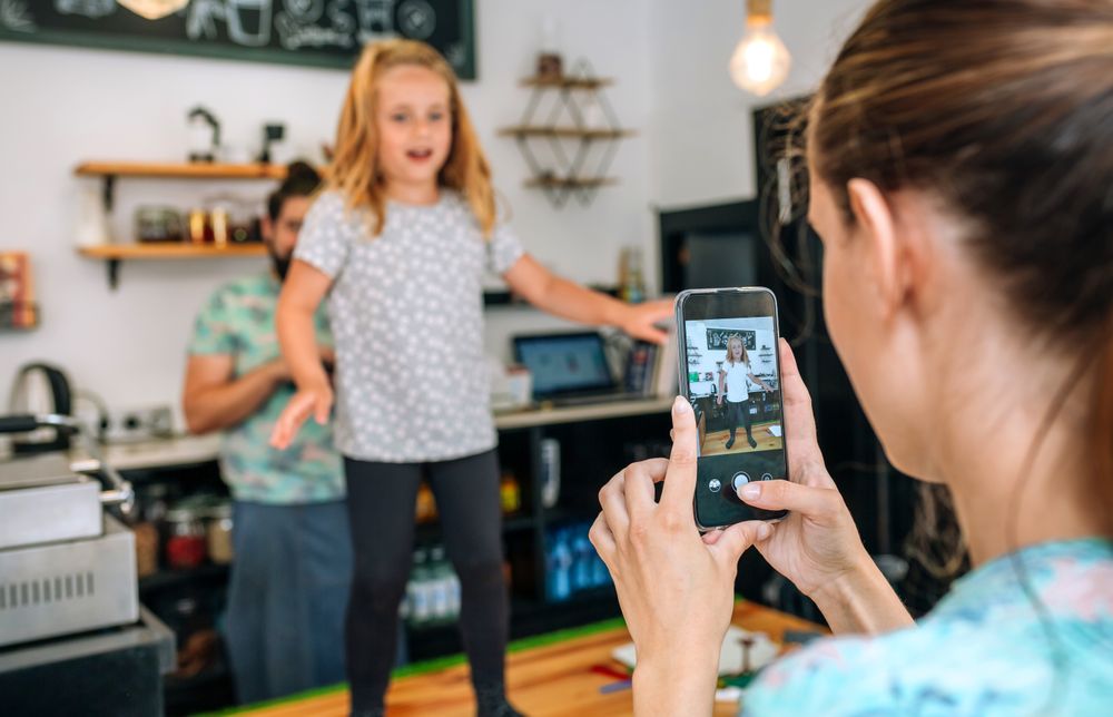 Mother recording her daughter dancing with the mobile while working in a coffee shop