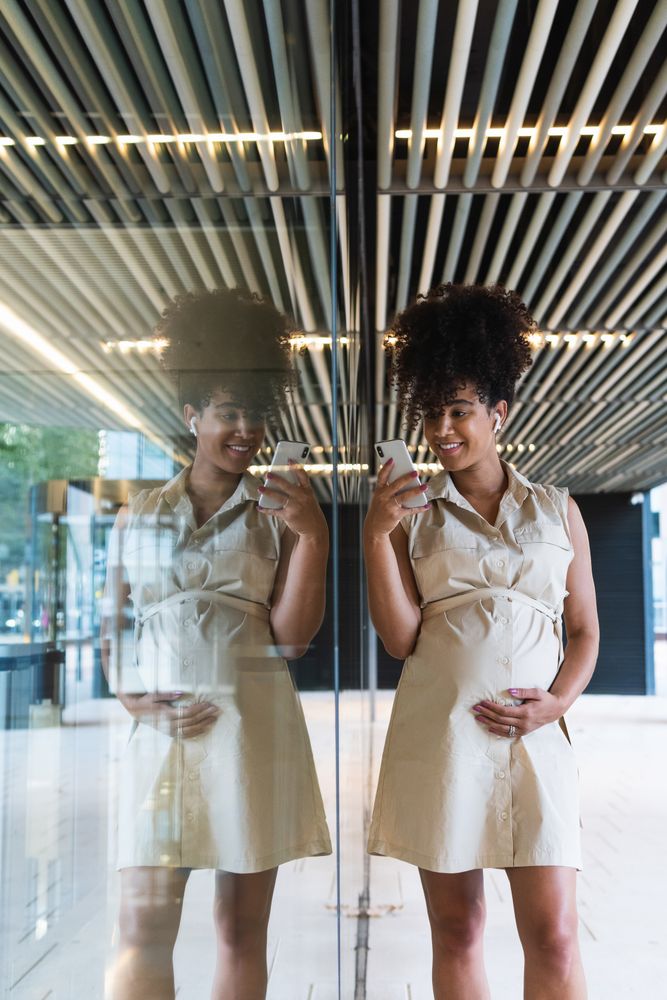 Smiling Black pregnant woman using smartphone and wireless earphones