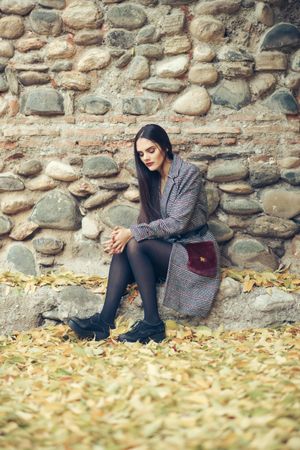 Female looking at camera in warm clothes sitting in front of rock wall in autumn time, vertical