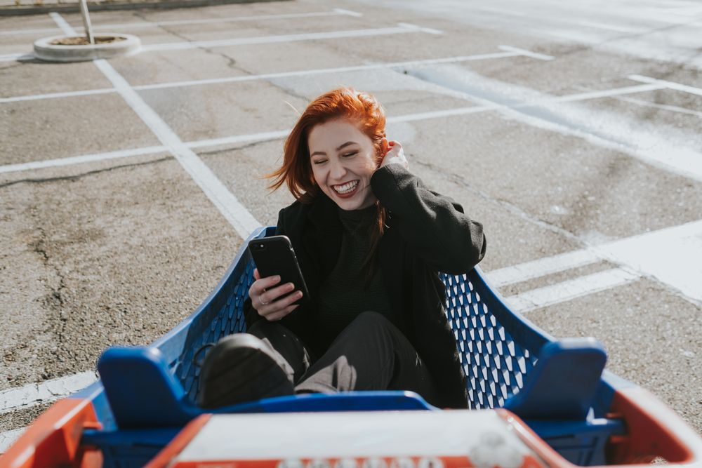 Smiling woman with smartphone in shopping trolley in parking lot