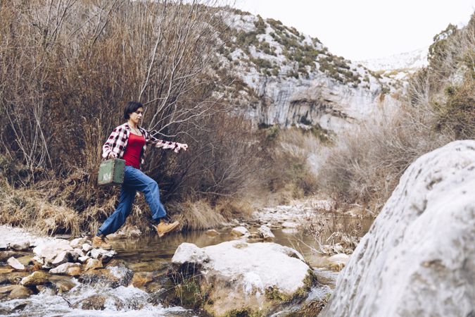 Woman with case jumping on rocks above stream