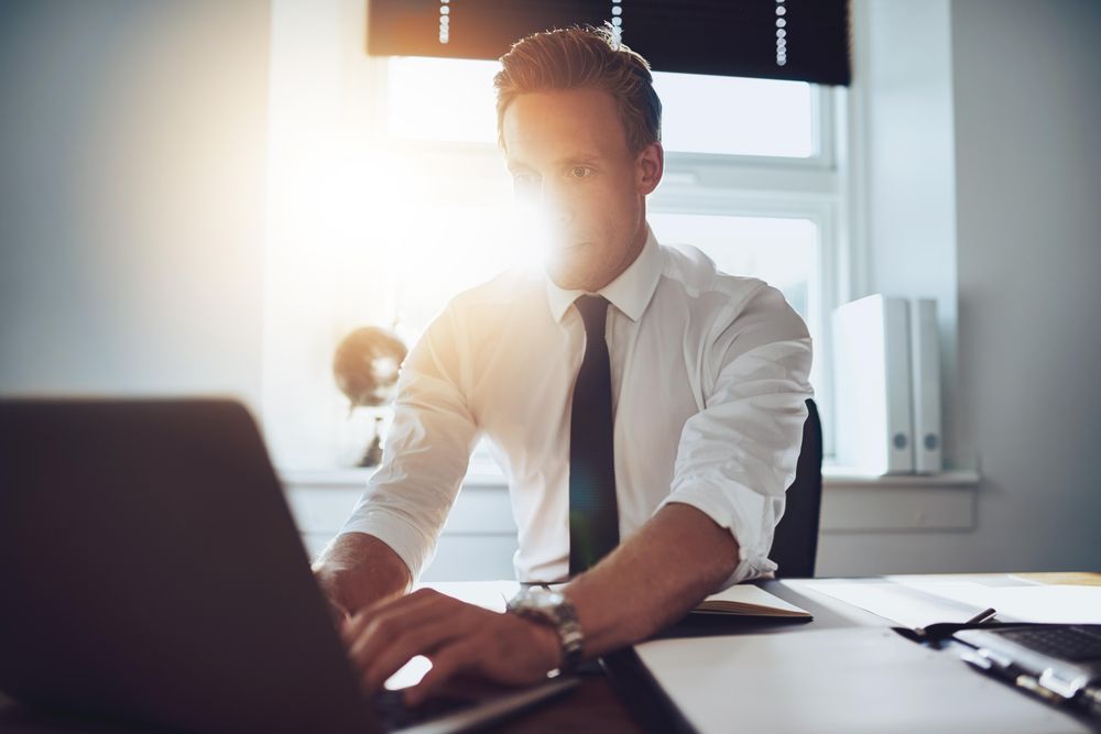 Man in shirt and tie sitting at desk typing at laptop in a sunny office
