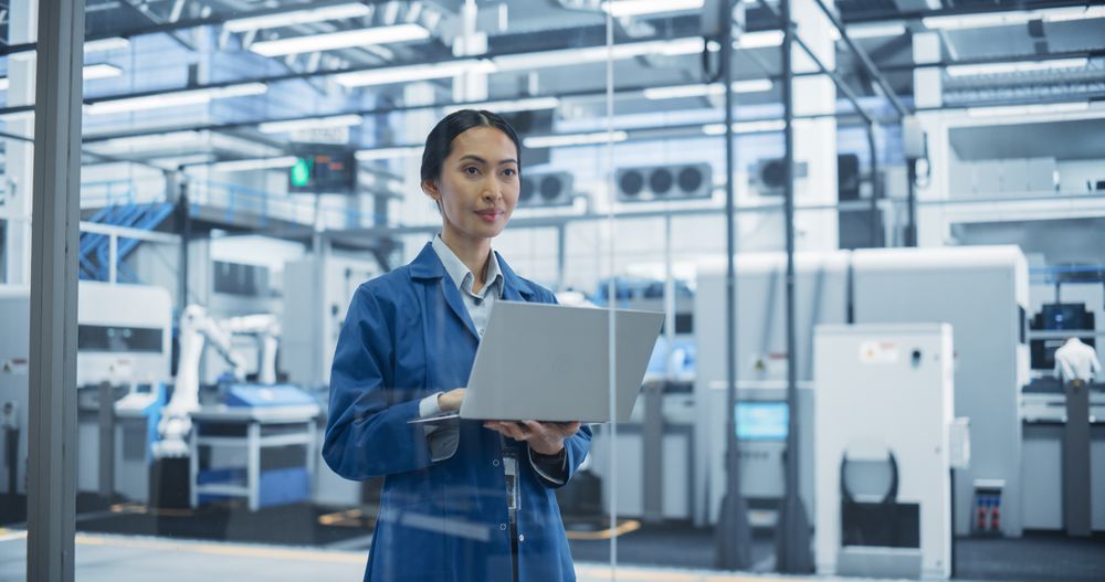 Electronics Manufacturing Technician in a Blue Coat Working on Laptop Computer at an Assembly Plant. Asian Female Specialist Developing, Maintaining, Setting Up Software for Automated Production