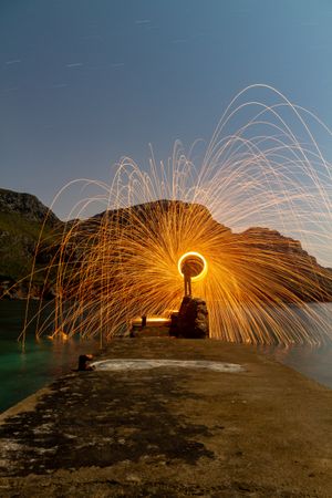Person with burning circle on stones between water near hills at night