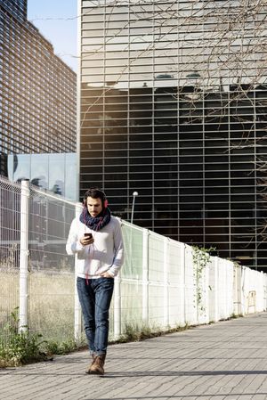 Young man in scarf walking past metallic fence and holding smartphone outdoors