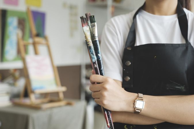 A close-up of the artist hands wearing an apron smeared with paint. Clutching many brushes and paintbrushes.