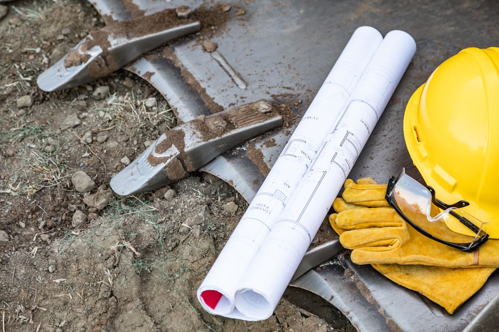 Technical Blueprints, Hardhat, Gloves and Protective Glasses Resting on Bulldozer Bucket Abstract