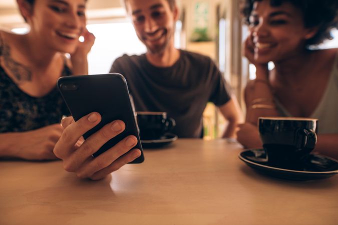 Three young people looking at mobile phone and smiling while sitting at restaurant table