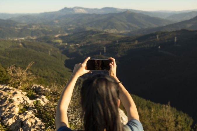A happy young woman takes pictures of the landscape with her smartphone from the top of a mountain