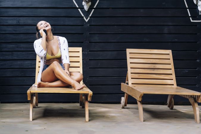 Delighted woman talking on phone while resting on deck chair