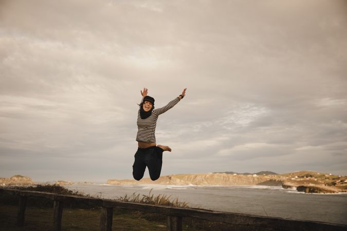 Young woman in hats having fun on seat on coast near waving sea and cloudy sky