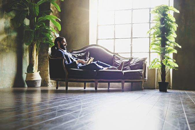 A Hispanic bearded businessman relaxes using a tablet lying on a sofa in the back of the office