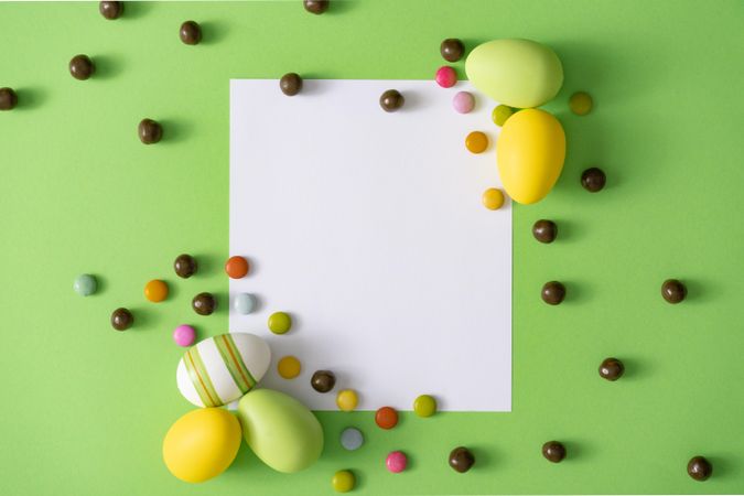 Easter eggs and chocolate pieces on green background with paper card square