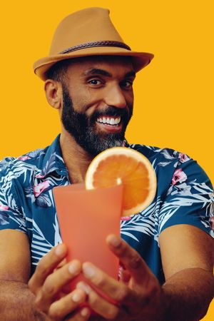 Happy male passing a cocktail to camera while wearing colorful shirt with yellow background