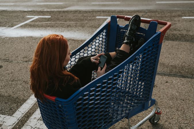 Woman with smartphone in shopping trolley in parking lot