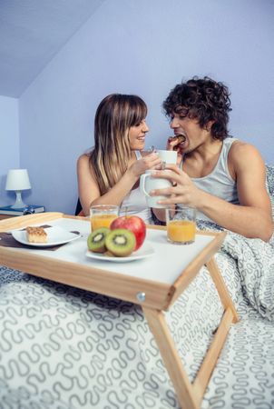 Young woman feeding happy man in bed breakfast
