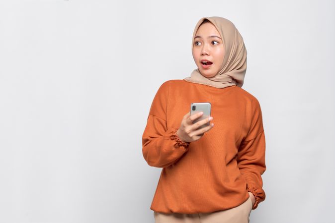 Surprised Muslim woman in headscarf and orange shirt with smart phone