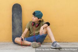 Front view of a teenager sitting on ground leaning on a yellow wall while holding a mobile phone 0WOnZO