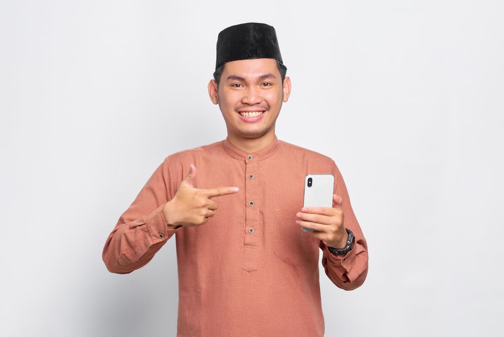 Happy Muslim man in kufi hat holding mobile phone and pointing at mobile phone