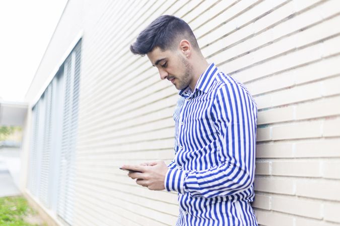 Side view of man leaning on house wall while using his smartphone