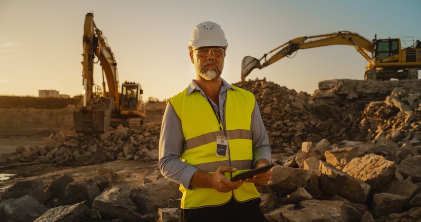 Successful Bearded Civil Engineer Wearing Protective Goggles And Looking At Camera On Construction Site on a Sunny Day While Heavy Machinery Working. Man Holding Tablet, Wearing Hard Hat, Safety Vest.