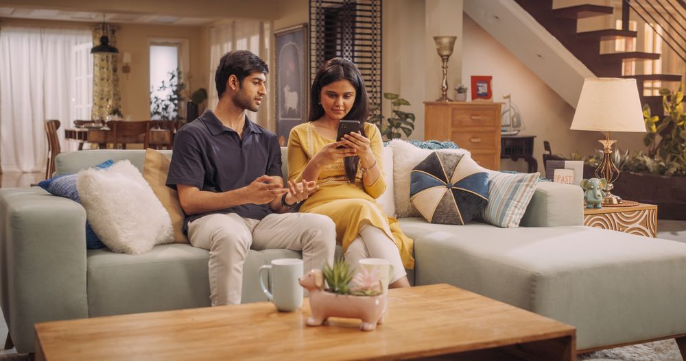 Young Indian Couple Using Internet On Smartphone, Sitting On The Sofa at Home, they Tease and Joke around.