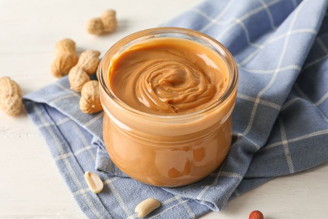 Glass jar with peanut butter, peanut and kitchen towel on wooden background, closeup and space for text