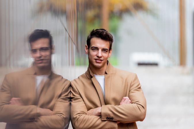 Man in fall coat smiling with arms crossed while leaning on reflective outdoor wall