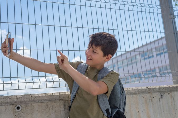 Smiling boy pointing at camera while taking selfie on smartphone outside of school yard