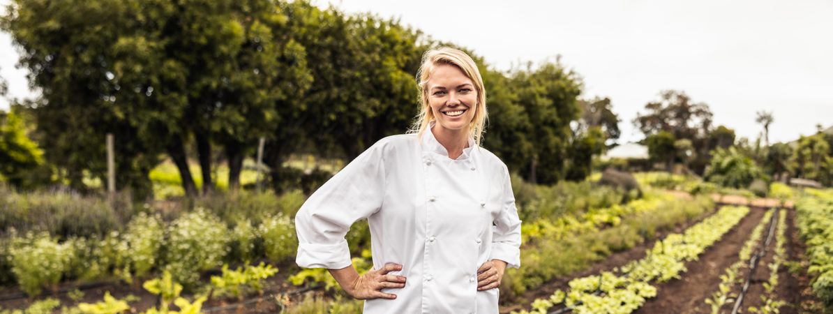 Cheerful chef smiling at the camera with her hands on her hips standing on a farm