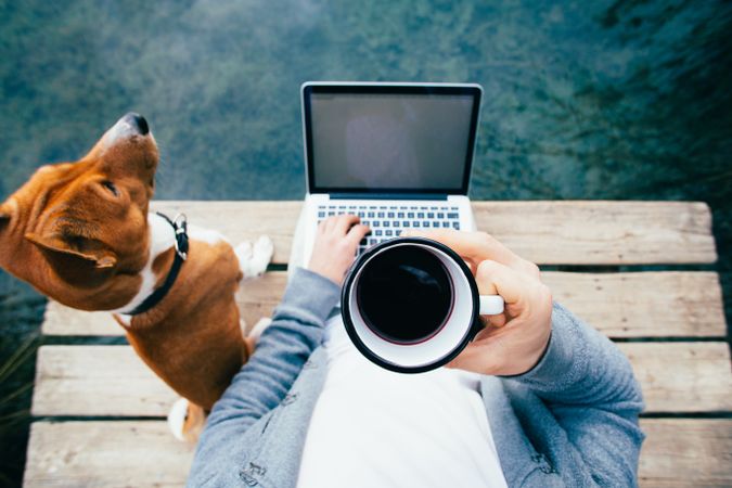 Man about to sip on coffee cup while working on the pier with pet dog