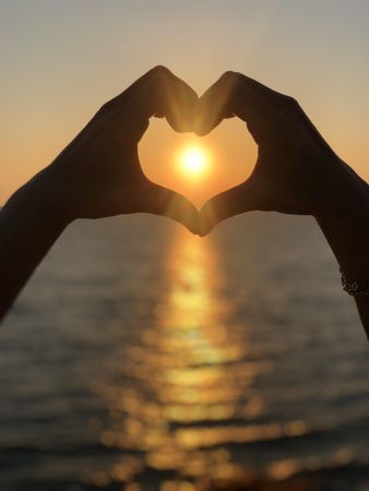 Persons hand forming heart near sea during sunset