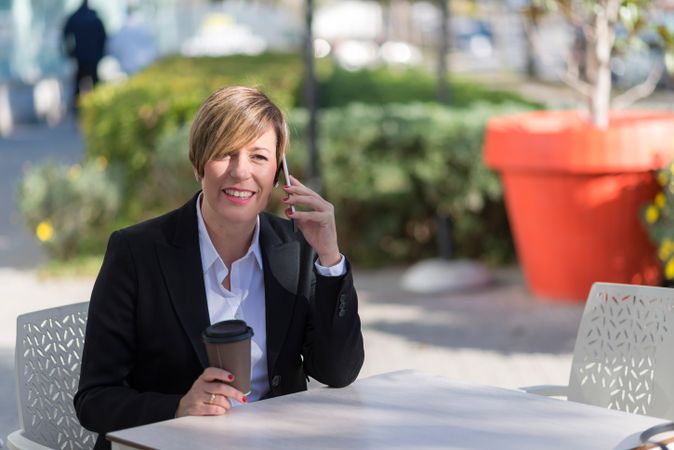 Smiling woman sitting on table outside with coffee speaking on cell phone