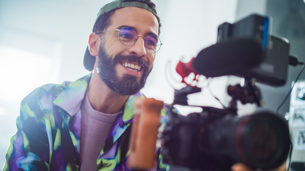 Smiling Young Latin Male Filmmaker Operating A Professional Camera In A Well-lit Studio, Capturing Engaging Content For A Modern Advertising Or Movie Production Project.