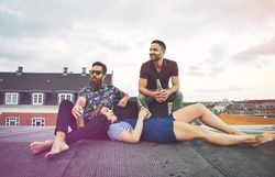 Two men and a woman lounging on a rooftop with beer
