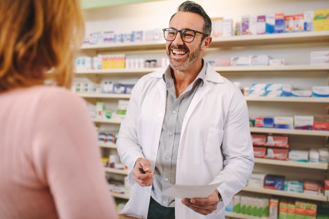 Smiling male pharmacist with prescription assisting a customer standing at the counter
