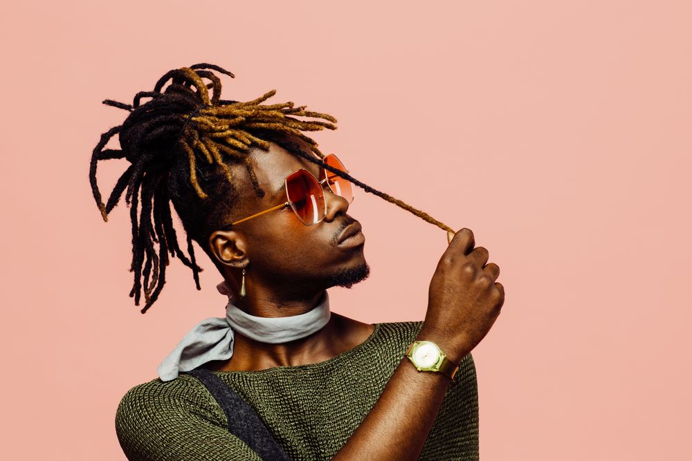Portrait of a young man in green with dreadlocks and blue sunglasses, isolated on pink