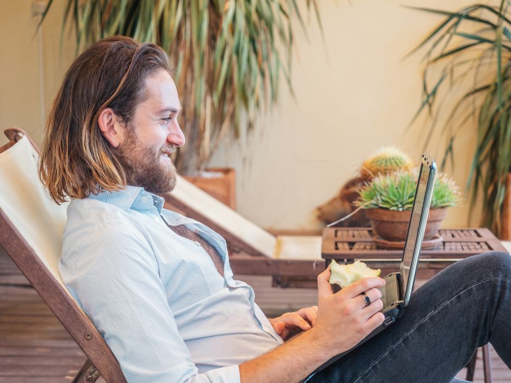Handsome male browsing laptop and enjoying apple on lounge in flat