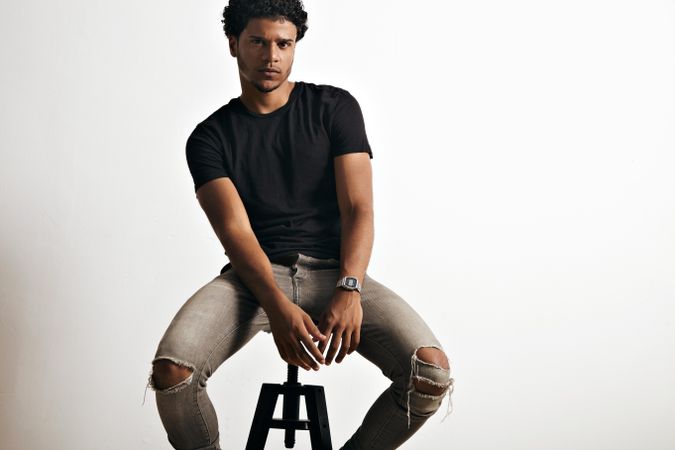Moody male model in dark clothes, ripped jeans sitting on stool in studio