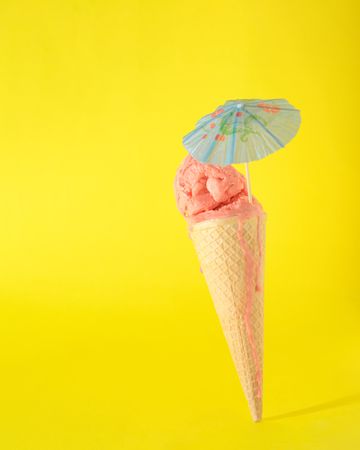 Cone with pink scoop of ice cream with cocktail parasol on yellow background