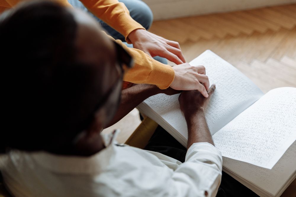 Person helping a visually challenged man to read a book using braille system