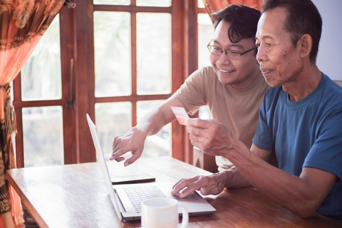 Happy adult son and older father pay online using laptop and credit card